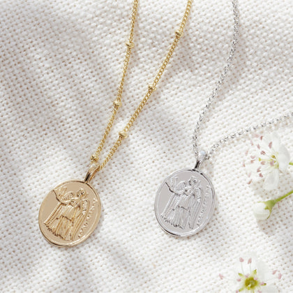 a gold athena coin charm on a gold bead chain laid out next to a silver athena coin charm on a silver belcher chain necklace. lying on a white fabric background